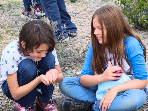 two girls sitting in desert one holding an insect the other looking on